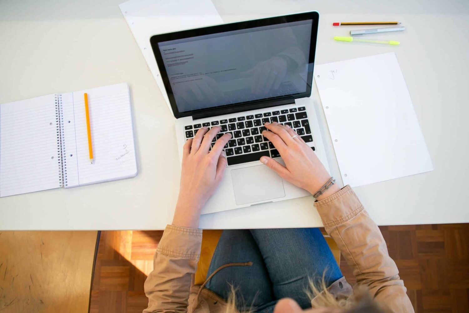 Vue de dessus d'une personne tapant sur un ordinateur portable, entourée de feuilles, d'un cahier et de stylos sur un bureau blanc