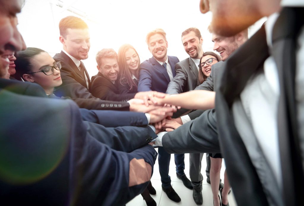 Groupe de professionnels en costume réunis en cercle, les mains jointes au centre dans un geste de cohésion d’équipe et d’esprit collaboratif