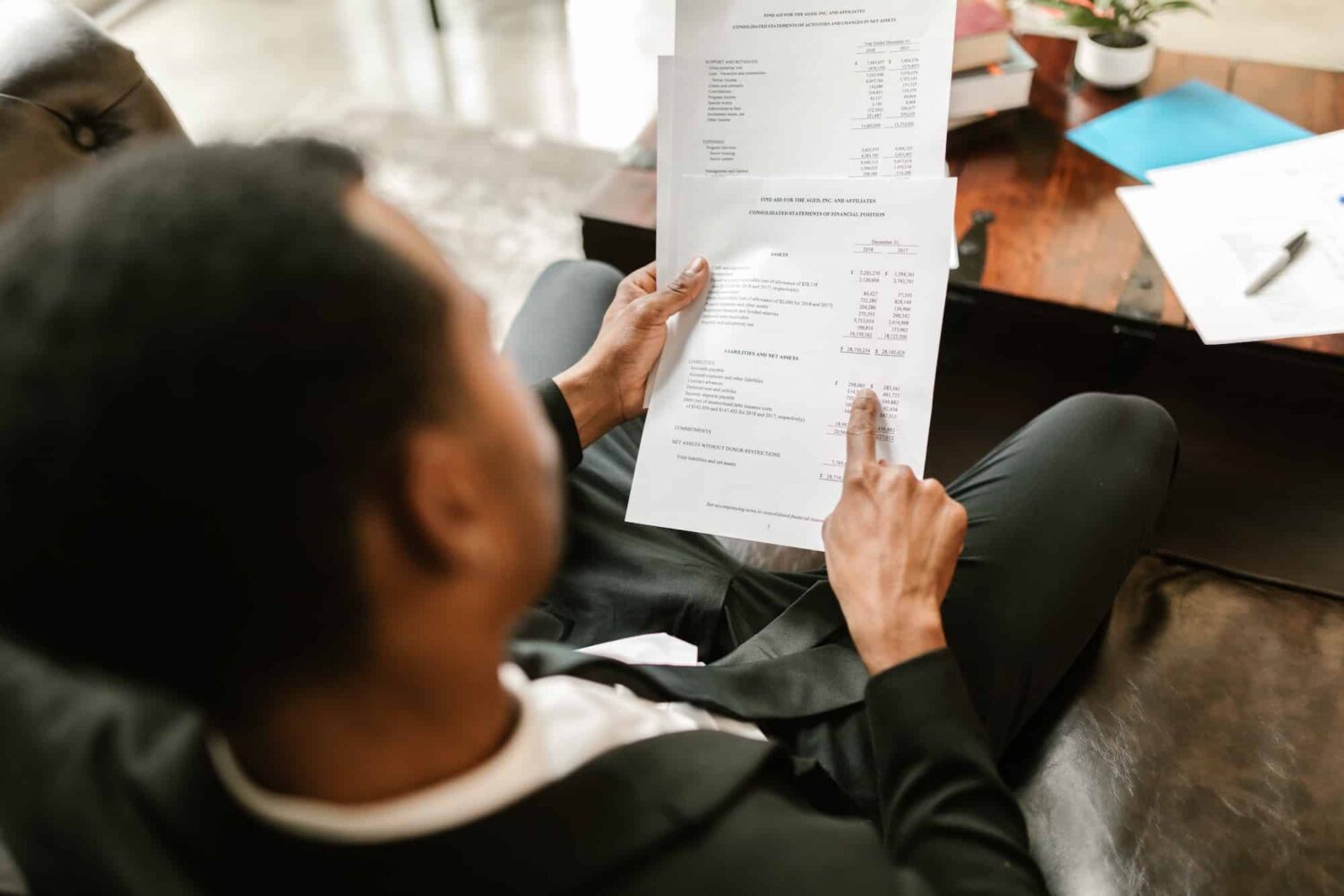 Homme en costume assis, consultant des documents financiers ou comptables, pointant une ligne sur une feuille contenant des chiffres.