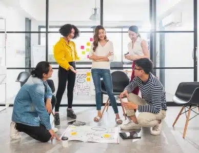 Groupe de jeunes adultes en workshop, discutant autour de plans au sol dans un espace de coworking lumineux