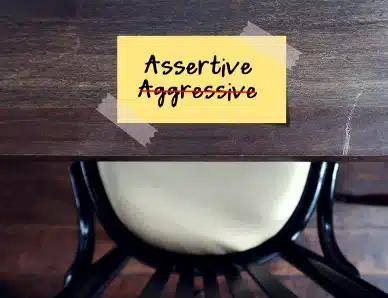 Note jaune collée sur un bureau avec les mots 'Assertive' et 'Aggressive' barré en rouge, illustrant la différence entre l'assertivité et l'agressivité.